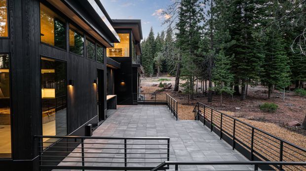 Exterior view of a contemporary multi-level home set among tall evergreen trees. The dark vertical-clad facade has extensive warm-lit glazing, projecting rooflines, and black metal-framed doors and windows. A broad grey-paved terrace and walkway with black horizontal-rail balustrades connect the building to the wooded site.
