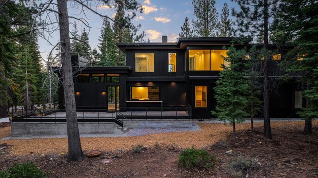 A modern two-storey dark-clad house set among tall evergreen trees in a forested setting. The building has a low, layered flat-roof form, extensive warm-lit glazing, black exterior cladding, concrete base and terrace edges, and a metal-railed outdoor deck. The composition is an exterior architectural photograph at dusk, with the warm interior lighting contrasting against the cool woodland surroundings.