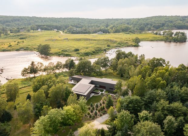 Aerial view of a modern, low-profile residence nestled among mature trees beside a broad body of water. The home has dark horizontal rooflines and a lighter projecting roof volume, with landscaped gardens and a winding driveway. Open grassy wetlands, wooded hills, and water surround the property.