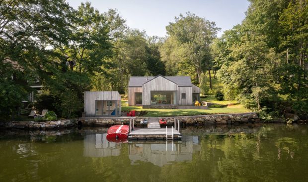 Exterior view of a contemporary lakeside residence set within dense woodland. The home features a simple gabled form, grey vertical cladding, large glazed openings, and a stone retaining edge along the water, with a small dock and red watercraft in the foreground.