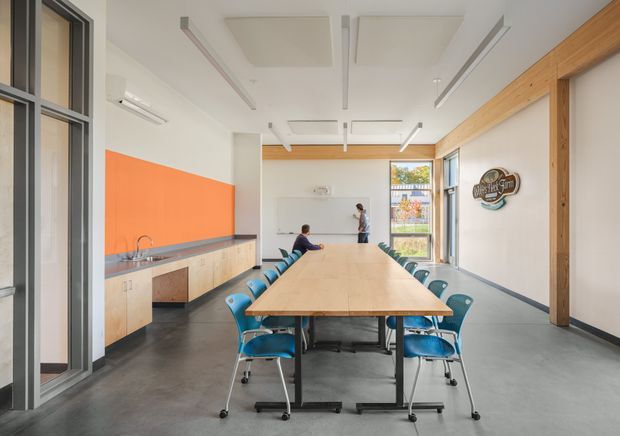 Bright contemporary communal meeting and dining room with a long timber table surrounded by teal chairs. The space features a polished grey concrete floor, white walls and ceiling, exposed timber structural posts and beams, an orange feature wall, and a compact kitchenette along the left side. Large glazed doors and windows bring in abundant daylight.