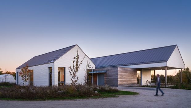 Exterior view of a contemporary single-storey building composed of interconnected pitched-roof volumes. The building has white horizontal cladding, dark standing-seam metal roofs, timber-toned horizontal wall cladding, tall glazed openings, and a covered recessed entry or passage. It sits in a landscaped rural or semi-rural setting at dusk.