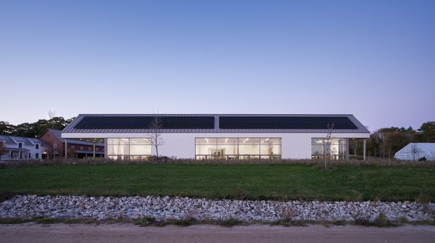 Wide exterior view of a low, elongated contemporary building at dusk. The building has a symmetrical shallow-pitched roof with dark photovoltaic panels, light-coloured walls, extensive illuminated floor-to-ceiling glazing, and a simple rectilinear facade. It sits behind a grassy verge and rock-lined drainage or shoreline edge, with neighbouring low buildings visible in the background.