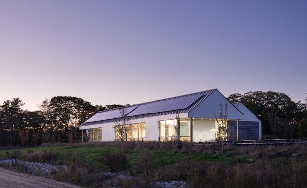 A single-storey contemporary rural house photographed at dusk. The building has a simple elongated form with a prominent pitched gable roof clad in grey metal, pale walls, large glazed openings, and a partially screened or louvred section on the right. It sits within a landscaped rural setting surrounded by trees and low planting.