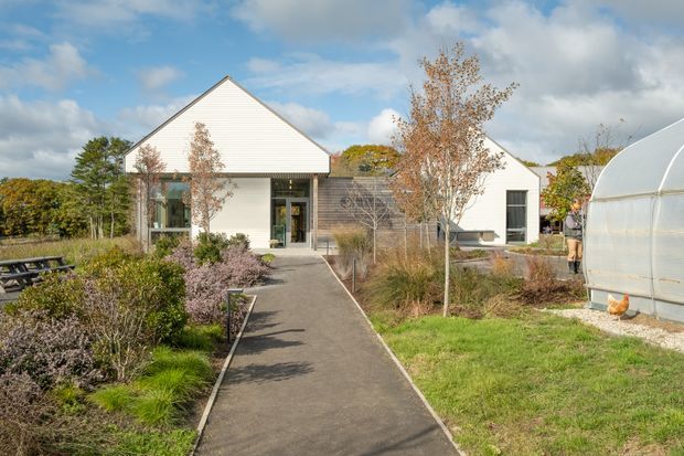 Exterior view of a low-rise contemporary building arranged around landscaped grounds, with a prominent white gabled volume, dark horizontal timber-like cladding, large glazed entry, and a paved pedestrian path leading to the entrance. The site includes ornamental planting, mature trees, outdoor seating, and a translucent greenhouse structure at right.