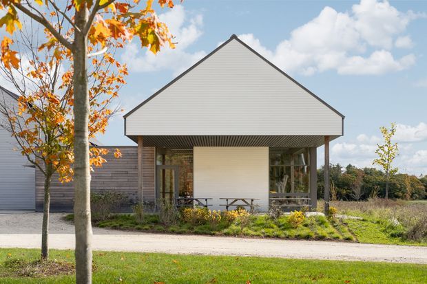 Front exterior view of a contemporary single-storey pavilion-like building with a steep symmetrical gable roof, light horizontal cladding, dark timber-framed glazing, and a covered entry or outdoor living space. The building sits within landscaped grounds with deciduous trees and a gravel walkway.