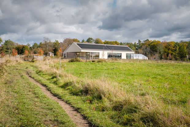 A single-storey contemporary rural house viewed across an open grassy field. The long, low building has a dark pitched roof fitted with solar panels, light-coloured exterior walls, extensive glazing, and a fenced garden or paddock area. Mature trees form the background, with a narrow dirt path leading toward the house under an overcast sky.