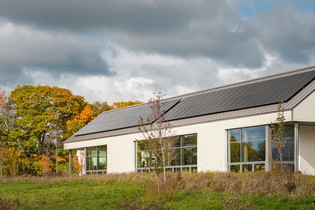 Exterior view of a low-rise contemporary building with pale horizontal weatherboard cladding, large glazed windows, and a dark pitched metal roof. The building sits in a grassy landscaped setting with autumn trees under an overcast sky.