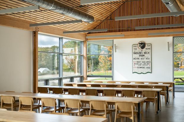 Bright communal dining hall with rows of timber tables and chairs, expansive floor-to-ceiling glazing, exposed timber structure and a slatted timber ceiling with exposed silver ventilation ducts. A large farm sign forms a wall feature, while landscaped surroundings are visible outside.