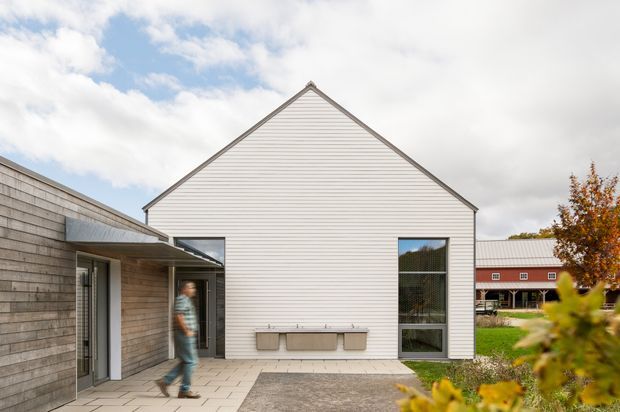 Exterior view of a contemporary gable-roofed building clad in light horizontal weatherboards, with a tall vertical window and paved courtyard entry. A lower timber-clad wing extends to the left, while a red barn-like structure is visible in the background. Autumn planting and a partly cloudy sky frame the scene.