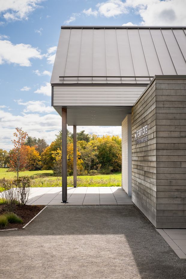 Exterior architectural photograph of a contemporary building entrance or covered walkway. The composition features a tall, simple metal-clad upper volume, a horizontal-panel facade, a deep projecting canopy supported by slender dark posts, and a textured horizontal-board wall with mounted signage. Paved paths, sparse planting, and a lawn with autumn trees surround the entrance under a bright partly cloudy sky.