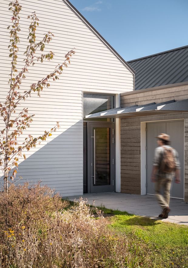 Exterior architectural photograph of a contemporary residential entrance. A white horizontal weatherboard-clad gabled volume meets a darker textured-clad lower wing with a charcoal standing-seam metal roof and projecting canopy. The entry includes a dark glazed timber or metal-framed door, an adjacent grey garage or utility door, concrete paving, and landscaped planting in the foreground. A blurred person is walking past the entrance.