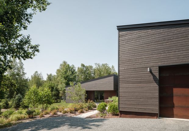 Exterior view of a contemporary low-rise house set within a wooded, landscaped site. The building has dark horizontal timber cladding, simple low-pitched or flat roof forms, large glazed openings, and a broad timber garage door. A gravel driveway and planted garden lead toward the recessed entry and living areas.