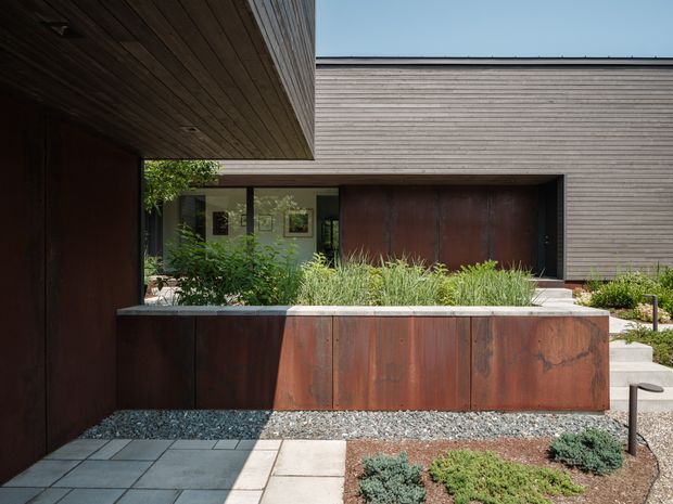 Modern single-storey residential facade and entry courtyard with dark weathered metal cladding, horizontal timber-like wall cladding, and a deep timber soffit. A raised planter clad in rust-toned metal separates the paved walkway from the recessed glazed entry, with minimalist gravel and drought-tolerant landscaping. The composition emphasizes crisp horizontal lines, recessed openings, and material contrast.