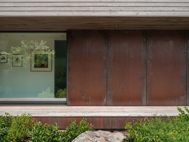 Close exterior view of a contemporary building facade featuring weathered dark timber cladding, a broad horizontal timber-framed window, and a deep timber soffit. The glazing reveals a softly painted interior with framed artwork, while low garden planting and a large stone sit along the base.