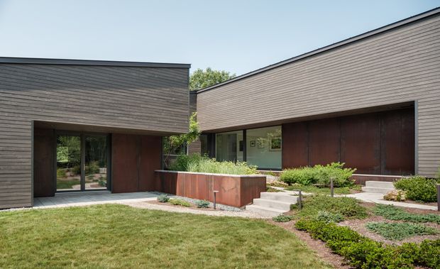 A low-slung modern residential building arranged as two connected wings around a landscaped courtyard. The exterior uses horizontal grey-brown timber cladding, dark-framed glazing, and large rust-toned recessed wall or door panels. Concrete steps, raised planter beds, gravel, clipped planting, and lawn create a structured contemporary landscape.