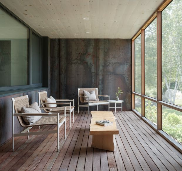 A serene enclosed outdoor lounge or sunroom with floor-to-ceiling glazing on one side, a dark weathered metal feature wall, timber plank flooring and ceiling, and slim-framed windows. Several woven lounge chairs with cushions surround a low sculptural timber coffee table, creating a calm indoor-outdoor sitting area overlooking greenery.