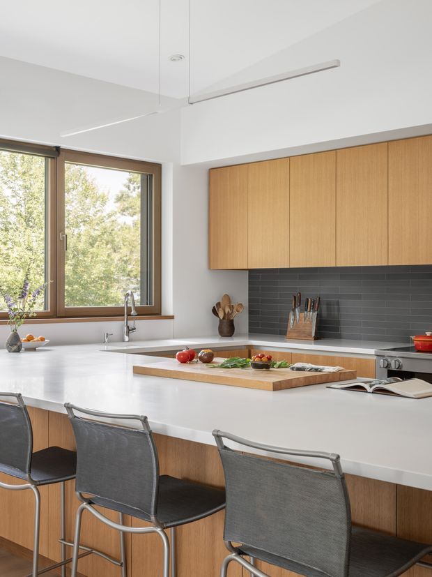 Bright, contemporary kitchen with a large white island and integrated seating, warm vertical-grain timber cabinetry, a dark grey horizontal-tile splashback, stainless-steel appliances and fixtures, and expansive timber-framed windows overlooking trees. Minimal linear pendant lighting and clean cabinetry create a refined, uncluttered interior.