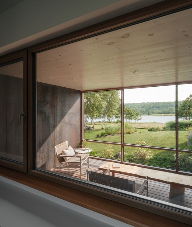Interior view looking through large floor-to-ceiling glazing onto a timber deck and landscaped waterfront setting. The space has a warm timber-lined ceiling, dark metal-framed sliding doors and windows, and a substantial dark textured feature wall. Outdoor lounge furniture is visible beyond the glass, with a calm lake and wooded shoreline in the distance.