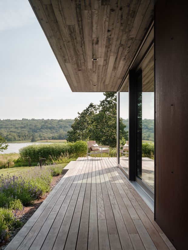 A covered timber deck or terrace extending along a modern home, with broad floor-to-ceiling glazing and dark vertical wall cladding. The deck and soffit use weathered timber boards, while a landscaped garden with lavender and seating overlooks a calm body of water and wooded hills. The composition emphasises the indoor-outdoor connection, wide overhang, and expansive view.