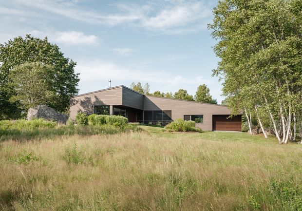 A low, single-storey contemporary house set within a natural grassy landscape and mature trees. The home has a simple angular roofline, dark timber-toned horizontal cladding, broad dark-framed glazing, and an integrated garage with a brown door. The restrained form and earthy exterior palette blend into the wooded setting.