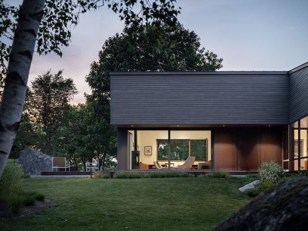 Twilight exterior view of a contemporary single-storey home set within a landscaped garden. The building has a dark horizontal-clad upper facade, broad floor-to-ceiling glazing, warm timber-toned wall panels, and a low-pitched roofline. Interior living spaces are visible through the windows, while mature trees, lawn, planting, and natural rock surround the house.