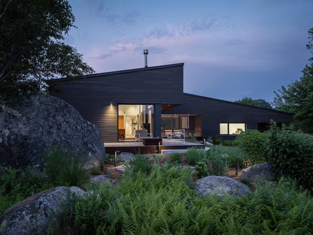 Exterior dusk view of a low-profile contemporary house set among natural boulders, ferns, and mature vegetation. The building has dark horizontal cladding, asymmetrical mono-pitched roof forms, broad glazed openings, a sheltered timber deck, and warm interior lighting.
