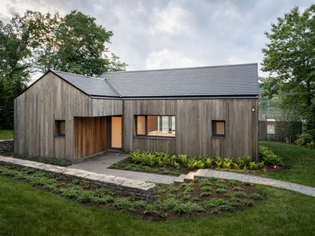 A compact modern single-storey house set within a landscaped, wooded site. The building has a simple gabled form, vertical weathered timber cladding, a dark grey standing-seam metal roof, dark-framed windows, and a recessed timber entry. Stone-edged paths and low planting lead to the entrance.