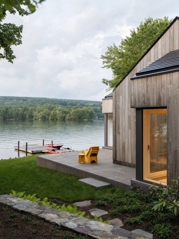 Exterior view of a modern lakeside house with a steep asymmetrical gabled roof, vertical weathered timber cladding, dark metal roof edges, and a warm timber-framed glazed entry door. A paved terrace leads toward a small dock and lake, with natural stone edging and landscaped planting in the foreground. The setting is wooded and waterfront, with an overcast but bright atmosphere.
