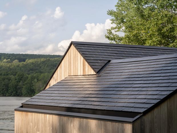 Close-up exterior view of a contemporary timber-clad building with a dark pitched gable roof covered in slate-like tiles. The roof has intersecting planes and a small projecting dormer or roof junction detail. A calm lake and wooded hills are visible in the background, with a mature tree at the right edge.