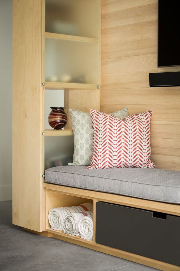 Close-up of a built-in timber window seat and storage wall. Pale wood joinery includes open shelving with frosted glass backing, a grey upholstered bench cushion, patterned cushions, towel storage in an open cubby, and dark lower cabinet fronts. A timber wall panel and partial wall-mounted television are visible at right.