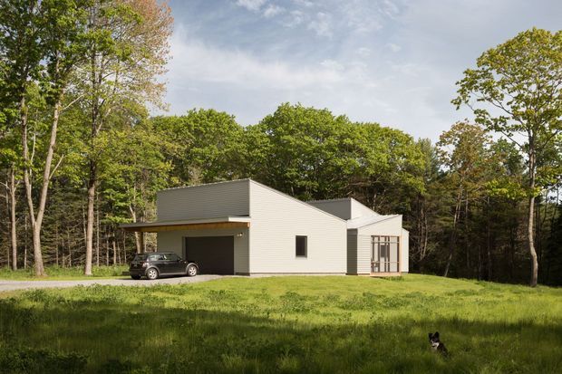 A minimalist single-storey contemporary house set within a wooded, rural landscape. The compact light-coloured building has a flat or very low-pitched roof, simple rectangular volumes, horizontal exterior cladding, a dark recessed garage or carport, and a glazed sunroom-like end section. A vehicle and small animal are present but are not part of the architectural subject.