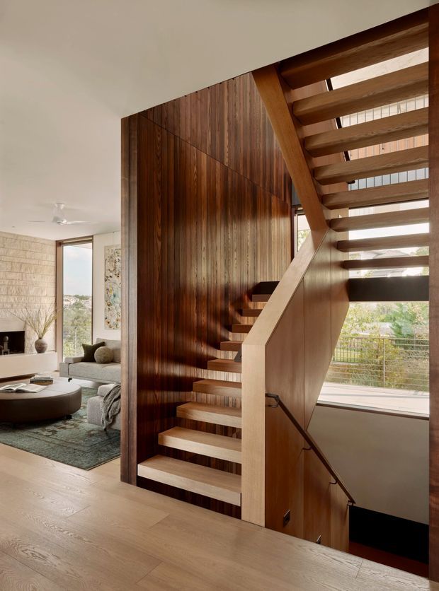 Interior view of a contemporary timber staircase rising beside a tall dark wood-panelled feature wall. The open-riser stair has warm timber treads, a pale solid side stringer, and a slim dark handrail descending to a lower level. Large windows bring in daylight and overlook greenery, while part of a softly furnished living room is visible to the left. Pale timber flooring continues through the space.