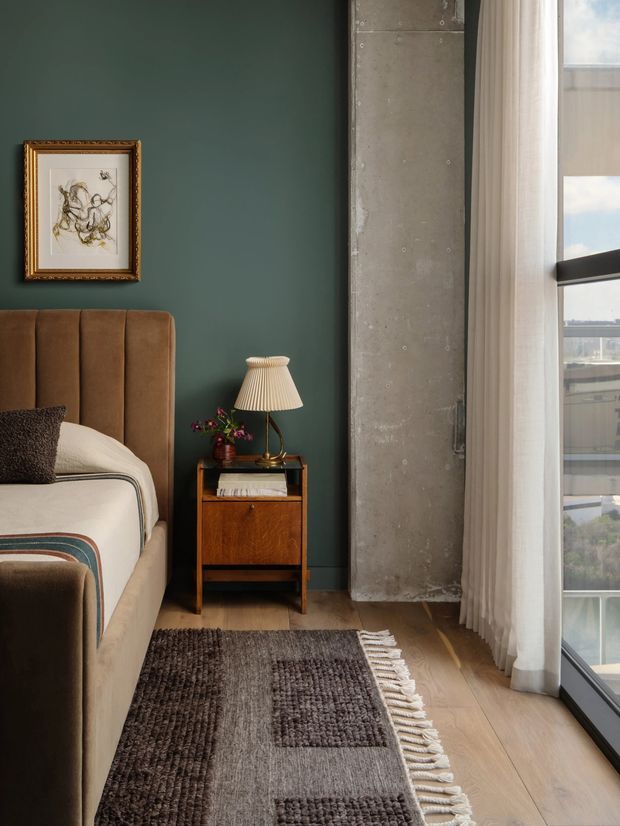 A warmly styled bedroom corner featuring a modern upholstered bed with a tall channel-tufted brown headboard, a dark green painted feature wall, framed artwork, and a vintage-style wooden bedside table with a pleated lamp. A raw concrete structural column and full-height window with sheer curtains add an industrial architectural contrast. Timber flooring and a textured neutral rug complete the composition.
