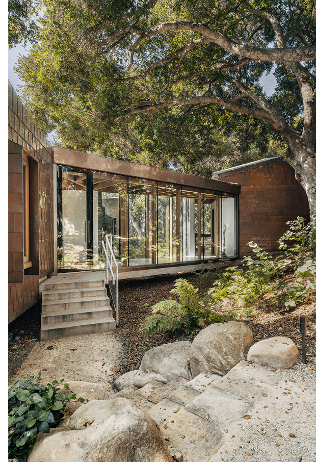 Exterior view of a low-profile modern building set within a wooded, landscaped site. A glazed connector or entry pavilion with extensive floor-to-ceiling windows links brown masonry volumes, with a flat or very low-pitched roof, exposed dark window framing, concrete steps and a metal handrail. Large established trees, ferns, rocks and mulch surround the building.