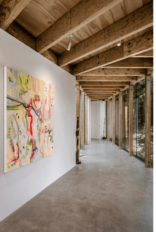 A long, gallery-like interior corridor with exposed timber framing and plywood ceiling panels. The concrete floor and white plaster wall create a restrained backdrop for a colourful abstract artwork, while a series of timber posts and beams line the glazed side overlooking trees. Small cylindrical pendant and recessed lights illuminate the passage.