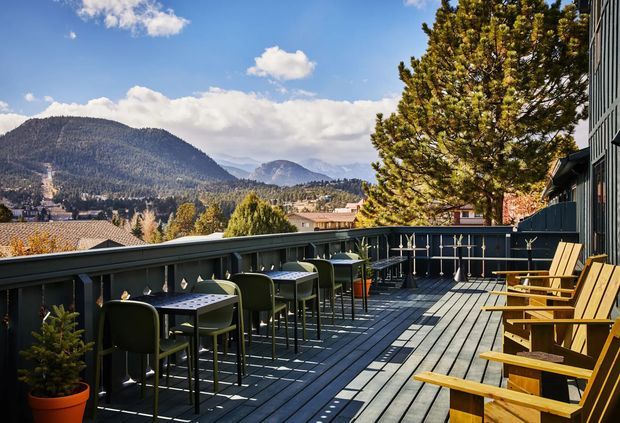 A spacious elevated outdoor terrace or balcony with dark timber decking, metal balustrades, bar-height tables and stools, and rows of wooden lounge chairs. The terrace overlooks a suburban mountain valley with forested hills under a bright blue sky. The setting appears suitable for outdoor dining and relaxation.
