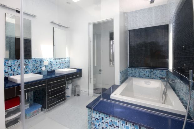 Bright contemporary bathroom with a double vanity, mirrored cabinetry, blue mosaic tile backsplash and bath surround, dark blue stone-like benchtops, a large built-in bathtub, and a glass-enclosed shower. White walls and pale flooring create a clean, high-contrast finish.
