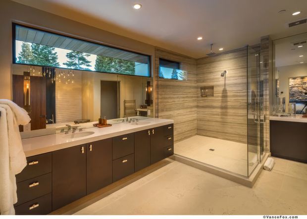 Spacious contemporary bathroom with a long dark-wood double vanity, white countertop and integrated sinks, large frameless glass shower enclosure, and full-height horizontally veined stone-look wall surfaces. Warm recessed lighting, clerestory windows, and mirrored cabinetry create a polished, luxurious atmosphere.