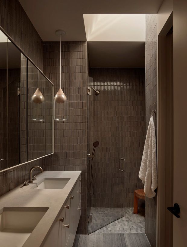 Compact contemporary bathroom with a walk-in shower, double vanity, large wall mirror, and sculptural pendant lights. The walls are clad in narrow vertically ridged dark brown tiles, contrasting with a pale stone vanity and mottled grey floor tiles. Warm, low-level lighting creates an intimate and refined atmosphere.