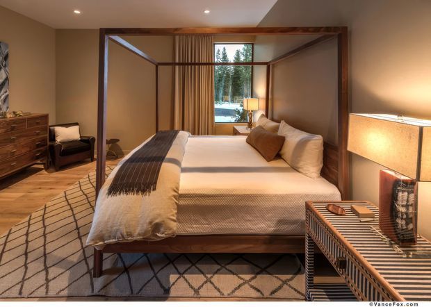 Warm, refined bedroom with a four-poster timber bed, white bedding, layered brown cushions, and a geometric area rug. The room features timber flooring, neutral painted walls, full-height curtains, a timber dresser, upholstered lounge chair, bedside lighting, and a large window overlooking trees. Built-in recessed ceiling lights and dark timber framing create a calm, sophisticated atmosphere.