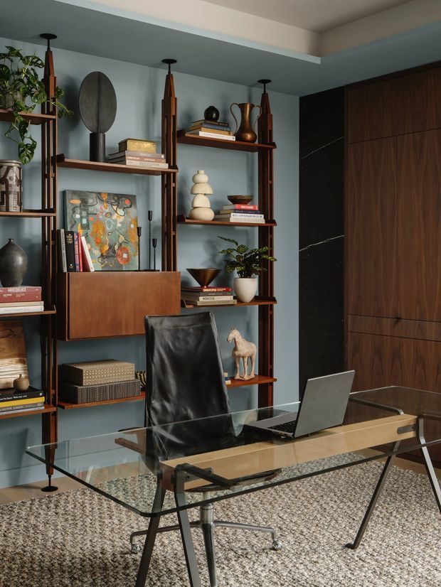 A refined home office with a glass-top desk, black leather office chair, open dark timber shelving filled with books and decorative objects, and a laptop. The room has blue-grey walls, dark walnut built-in cabinetry, a textured neutral carpet, and a sophisticated mid-century-inspired atmosphere.