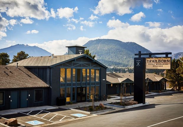 Exterior view of the Trailborn Rocky Mountains hotel complex, featuring a dark timber-clad, pitched-roof building with a prominent glazed entrance and a tall branded roadside sign. Paved parking and landscaped beds sit in front, with dramatic forested mountains visible behind the property.