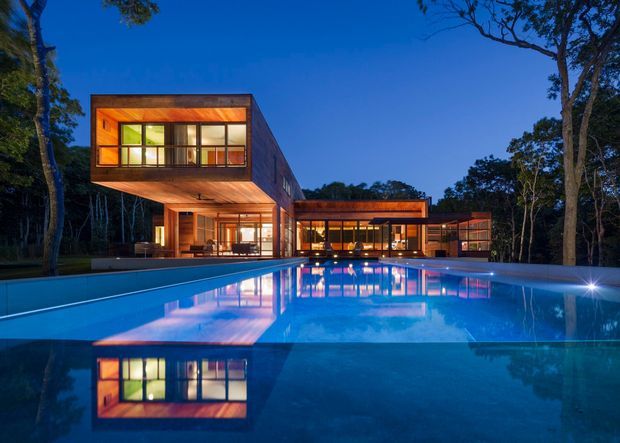 Twilight exterior view of a modern multi-wing residence set among trees, with warm timber-clad volumes, extensive glazing, projecting upper-level forms, and a large illuminated swimming pool reflecting the building.