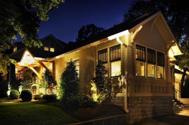 Night-time exterior view of a warmly illuminated traditional single-storey house, featuring gabled roof forms, cream weatherboard cladding, stone-look masonry at the base, dark-framed multi-pane windows, timber brackets and exposed decorative gable detailing. Landscaped garden planting and pathway surround the facade.