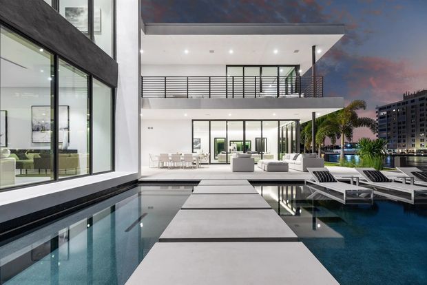 A contemporary multi-level waterfront residence photographed at dusk, centred on a long reflective swimming pool with broad stepping slabs leading toward the house. The home features crisp white rendered walls, extensive floor-to-ceiling glazing in dark frames, projecting balconies with horizontal metal balustrades, a flat roof, and an open outdoor entertaining area with lounge furniture and poolside loungers. Interior living and dining spaces are visible through the glazing, with city buildings and water beyond.