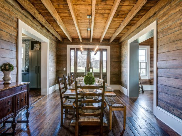 A rustic dining room in a timber-lined interior, centred on a long wooden dining table with ladder-back chairs and place settings. Exposed timber ceiling beams, weathered horizontal wall boards, dark timber flooring, and white-trimmed openings define the room. Glazed doors at the rear open toward an outdoor view, while adjacent rooms are visible through the side openings.