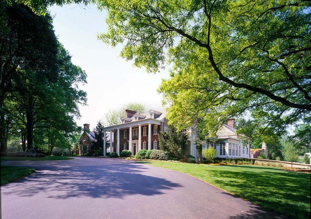 A grand traditional two-storey residence viewed from a broad paved driveway, set within mature landscaped grounds. The façade features red brick, white classical columns and trim, tall multi-pane windows, prominent gabled and hipped roof forms, dormer windows, and multiple chimneys. Large established trees frame the property.
