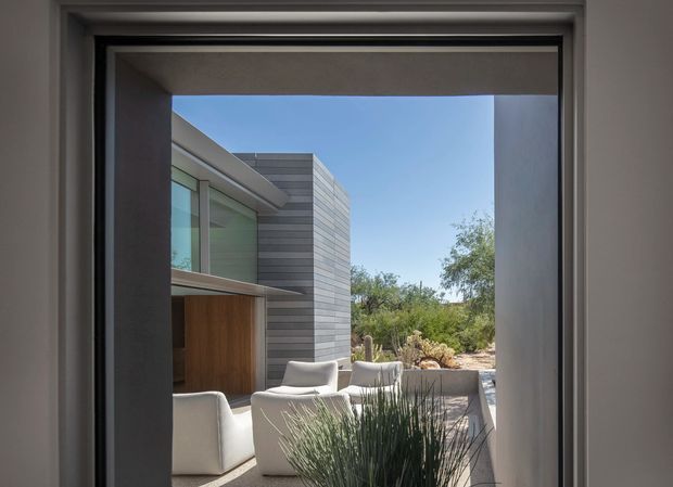 View from inside through a large framed opening toward a modern courtyard terrace. The scene shows a rectilinear building with horizontal grey cladding, extensive glazing, a timber-toned entry or wall panel, pale outdoor lounge seating, and drought-tolerant landscaping under a clear sky. The image is dominated by the architectural exterior and courtyard beyond the interior threshold.