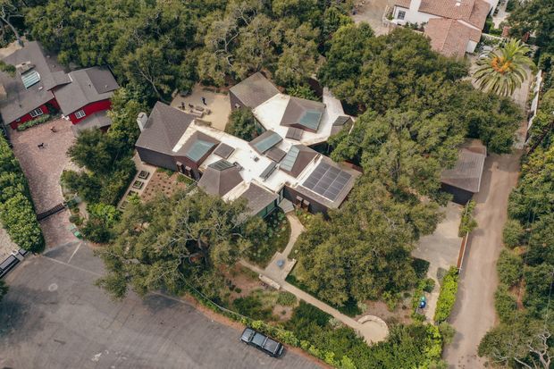 Aerial view of a large, low-rise residential property surrounded by dense mature trees and landscaped garden areas. The home has multiple interconnected pitched roof volumes, several skylights, and a prominent roof-mounted solar panel array. A driveway and neighbouring homes are visible around the wooded suburban site.